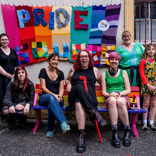 Jody Toomey sits on a pride bench at Newtown square, there are other people on the seat with her and standing at either side of the bench. In the background a crochet yarn bomb sign reads PRIDE SQUARE, everyone is smiling